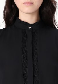 Blusa nera con colletto arricciato, un bottone decorativo e dettagli in pizzo verticale sulla parte frontale. Tessuto morbido con una texture liscia.