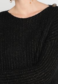 Svart stickad tröja med ribbad textur, lös passform och vid halsringning. Har en knappdetalj på axeln.