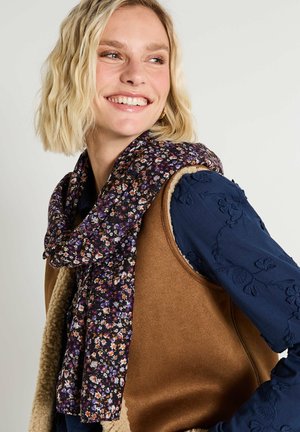 Écharpe florale avec un fond violet foncé et des fleurs multicolores, posée lâchement autour du cou d'un mannequin, contrastant avec un gilet marron.