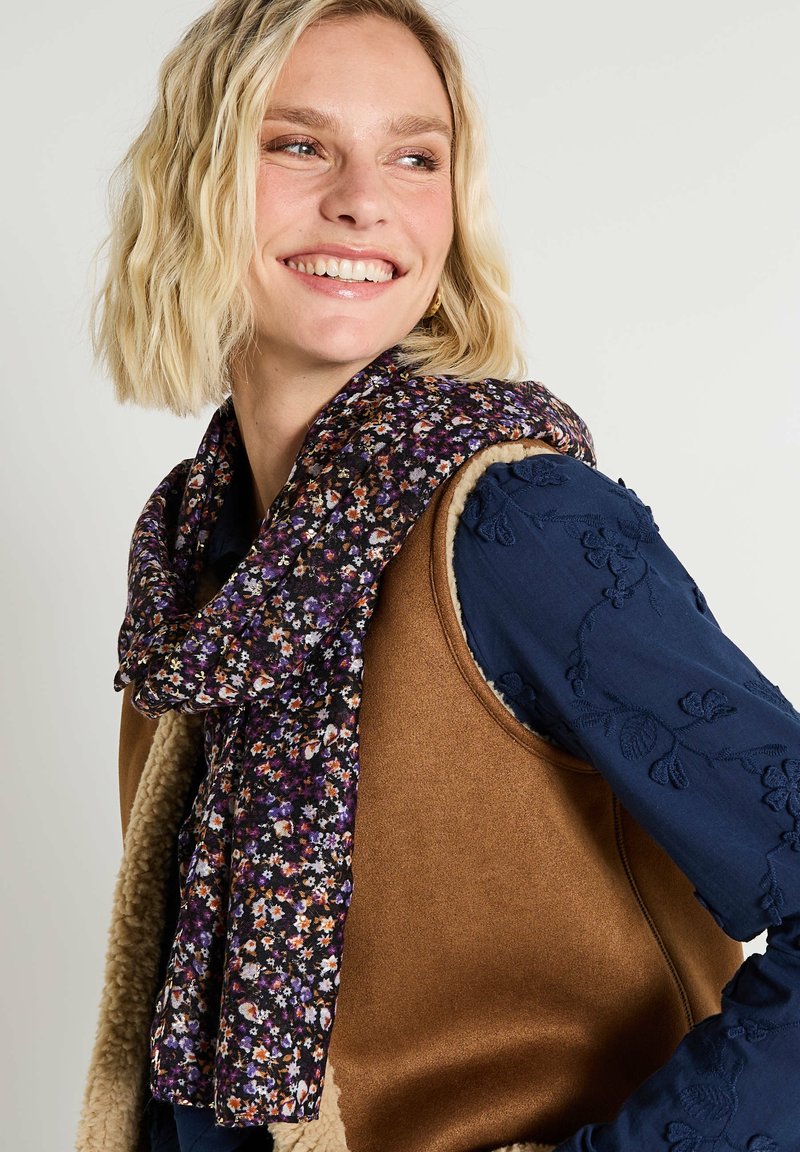 Écharpe florale avec un fond violet foncé et des fleurs multicolores, posée lâchement autour du cou d'un mannequin, contrastant avec un gilet marron.