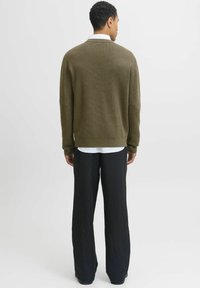 Vīrietis ar muguru pret skatītāju, uzvilcis olīvu zaļu džemperi, apakšā balta krekla piegriezums un melni svītroti plata kāju bikses, pret vienkāršu fonu.