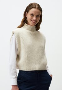 Pull en tricot sans manches couleur crème avec un col côtelé haut, en tissu texturé, porté sur une chemise blanche à manches longues, associé à un pantalon sombre.