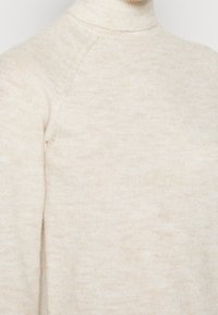 Cremfarbener Strickpullover mit Stehkragen und langen Ärmeln, der aus einem weichen Textil mit subtilen Melange-Muster besteht.