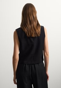 Top negro sin mangas con cuello alto, con una textura suave, detalle de costura en la parte posterior y un largo cropped, combinado con pantalones negros holgados.