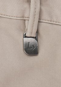 Zbliżenie na beżową tkaninę pokazuje metalowy zamek z logo. Tekstura jest pleciona, a zamek jest prostokątny z gładkimi krawędziami.