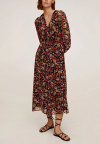 Femme portant une longue robe florale noire avec des fleurs rouges et orange et des sandales lacées noires, se tenant devant un fond uni.