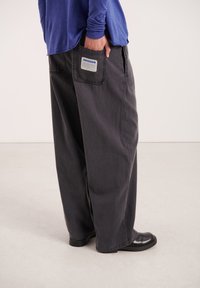 Voľné čierne džínsy s textúrovaným povrchom, s označením na zadnom vrecku a efektom opotrebovania, kombinované s modrou košeľou s dlhým rukávom a čiernymi topánkami.