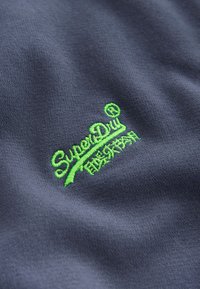 Tela azul oscuro con el logotipo "SuperDry" bordado en verde brillante y caracteres japoneses debajo.