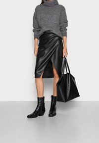 Person som bär en grå stickad polotröja, svart läder omslagskjol, svarta ankle boots, och håller en stor svart läder tote-väska.