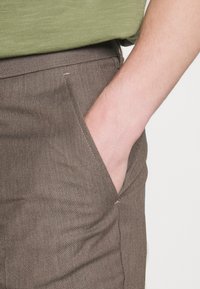 Pantalon marron tissé avec une finition texturée, doté d'une poche latérale dans laquelle une main est insérée. La ceinture est fine, et un haut vert est partiellement visible.