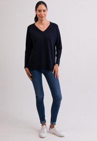 Svetle modrý svetr s výstřihem do V z měkké látky, volný střih. V kombinaci s přiléhavými modrými džíny a bílými teniskami, postavený proti světlému pozadí.