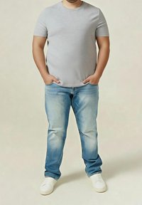 Mężczyzna stojący ze złożonymi rękami w kieszeniach, ubrany w jasnoszary t-shirt, wyblakłe niebieskie dżinsy oraz białe trampki na jednolitym tle.