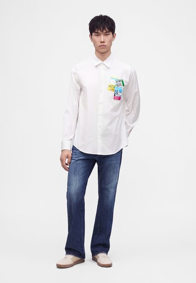Jeune homme en chemise blanche boutonnée avec des patchs colorés sur la poche poitrine, jean bleu et baskets beiges, debout sur fond blanc.