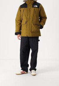 Kurtka puchowa w oliwkowym kolorze z czarnymi akcentami, dwa przednie kieszenie i kaptur z sztucznego futra. Połączona z ciemnymi dżinsami i kremowymi butami sportowymi w czerwone paski.