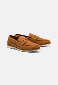 Kellot nahka loafersu tummasta nahasta, keveä valkoinen kumipohja ja koristeellinen nauha varpaankannen yli. Timberland-logo sivussa.