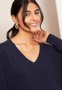 Granatowy sweter w serek o miękkim, teksturowanym splocie, z długimi rękawami i prążkowanymi mankietami. Modelka ma na sobie złote kolczyki w kształcie kół oraz gładko ułożone włosy.