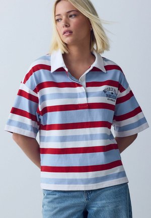 Kobieta w polo w czerwone, białe i jasnoniebieskie paski z napisem „Beverly Hills Padel Club Los Angeles”, w połączeniu z niebieskimi dżinsami.