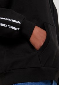 Sudadera negra con capucha que presenta un bolsillo frontal, puños acanalados y rayas de marca blancas a lo largo de la manga para mayor contraste. Tejido suave y texturizado.
