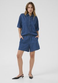 Denim krekls ar īsām piedurknēm un priekšpuses pogām, saskaņots ar atbilstošiem džinsu šortiem, abi vienā cietā zilo krāsā. Melni siksnas augstpapēžu apavi.