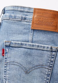 Jeans en denim bleu clair avec un patch en cuir portant un logo en relief, une étiquette rouge sur le côté et des détails de couture classiques sur les poches arrière.