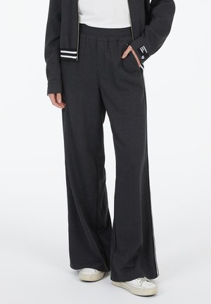 Person wearing dark gray wide-leg pants with white side stripes, white sneakers, and a black jacket with striped cuffs, hands resting in pockets.