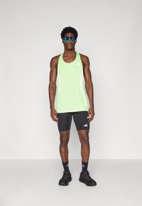 Limoengroene tanktop met witte accenten, gecombineerd met zwarte shorts. Het model draagt zwarte schoenen en blauwe reflecterende zonnebril.