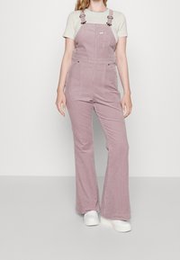 Rosa kordoveralls med justerbara axelremmar, utsvängda ben och framfickor, kombinerade med en ljusrandig t-shirt och vita sneakers.