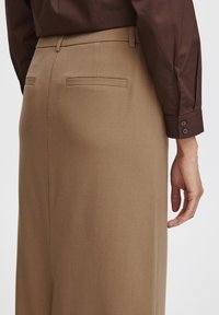 Bruna skräddarsydda byxor har en jämn textur, två bakfickor och diskret sömnad. En mörkbrun skjorta är delvis synlig.