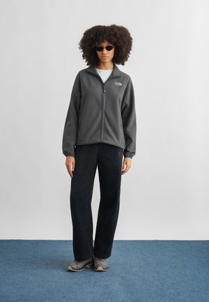 Γυναίκα με μαλλιά σε μπούκλες και γυαλιά ηλίου φοράει γκρι φλις ζακέτα με φερμουάρ, ανοιχτόχρωμο πουκάμισο, μαύρο παντελόνι και γκρι αθλητικά παπούτσια, στέκεται πάνω σε μπλε χαλί.