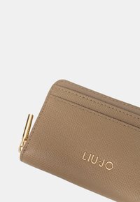 Cartera de cuero texturizado marrón con logo en tono dorado y tirador de cremallera. El diseño delgado cuenta con compartimentos para tarjetas y detalles de costura limpios.