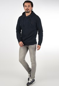 Marineblauwe hoodie met verstelbare trekkoorden, kangoeroezak en logopatch. Gecombineerd met lichtgrijze slim-fit jeans en donkere sneakers.