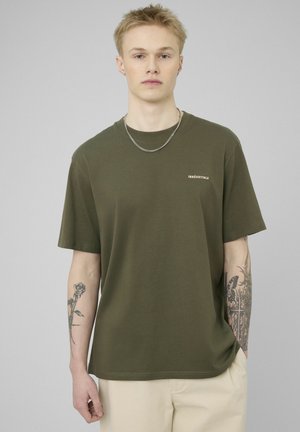 Jeune homme aux cheveux blonds courts portant un t-shirt vert olive avec le texte « IRRÉSISTIBLE », un pantalon beige, une chaîne en argent et des tatouages floraux sur les deux bras.