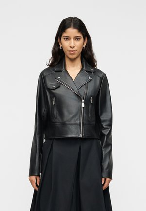 Femeie cu păr lung și închis la culoare poartă o jachetă de piele neagră, scurtă, cu buzunare cu fermoar și o fustă neagră plisată, stând în picioare pe un fundal simplu.