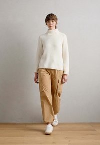 Maglione bianco a costine con collo alto, pantaloni cargo beige con tasche laterali e sneakers bianche. Il modello è in piedi su un pavimento in legno contro uno sfondo neutro.