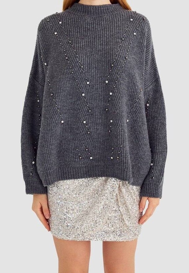 Pull en laine gris avec de petites perles dispersées, associé à une jupe courte argentée à sequins, porté par une personne debout.