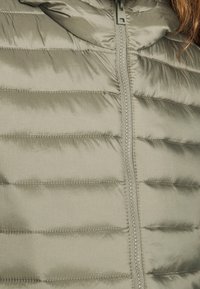 Olivově zelená péřová bunda s vertikálními švy, zipovým uzávěrem a vysokým límcem. Hladký, lesklý materiál s kontrastním prošíváním.