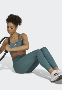 Teal sports bra and matching leggings made from stretchy fabric, with a fitted design. The athlete holds a black exercise hoop. White sneakers.