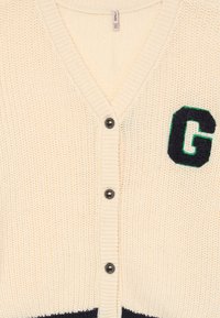 Beige stickad cardigan med V-hals, svarta knappar och en grön och marinblå bokstav "G"-applikation på vänster sida. Strukturerad tyg.