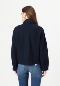 Giacca in misto lana blu navy con colletto strutturato, spalle scese e vestibilità leggermente accorciata, caratterizzata da un tessuto testurizzato e cuciture minime.