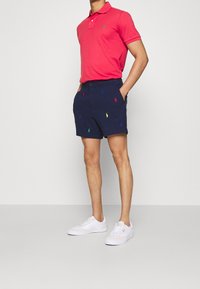 Camisa polo rosa con un logotipo bordado, pantalones cortos azul marino con logotipos multicolores y zapatillas blancas. El modelo está de pie con las manos en los bolsillos.