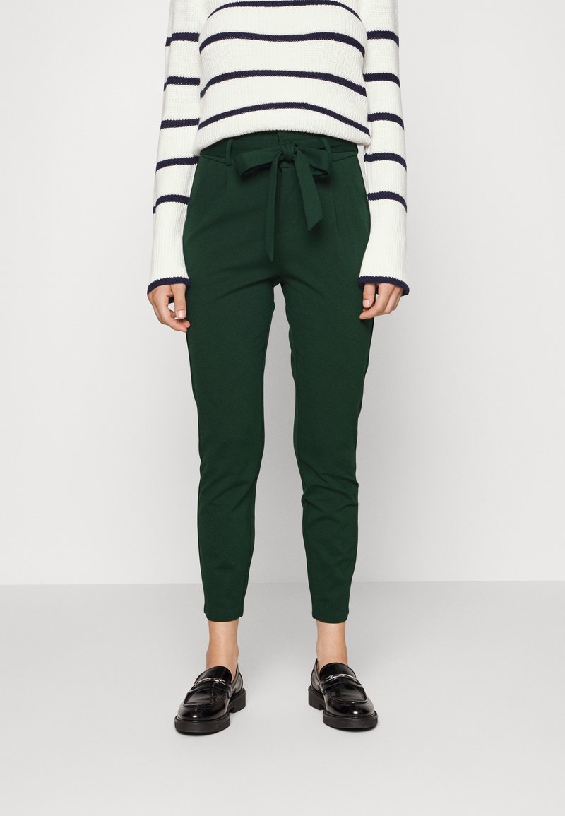 Femme portant un pantalon taille haute vert foncé avec un lien à la taille, un pull blanc à rayures horizontales noires et des mocassins noirs, debout sur un fond uni.
