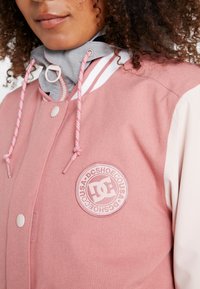 Kobieta w różowo-kremowej kurtce DC Shoes z szarą bluzą z kapturem pod spodem, z widocznym logo DC Shoes na klatce piersiowej.