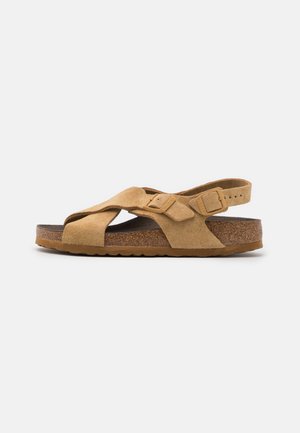 Sandalia de ante beige con diseño de tiras cruzadas, hebilla ajustable y plantilla de corcho. Suela plana y texturizada para mayor tracción. Vista lateral.