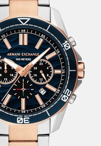 Armani Exchangei kronograafkellaga, millel on sinine põhjapind, roosakuldne aktsent ja hõbedane roostevabast terasest korpus. Omab kolme alamkohta ja kuupäeva näidikut.