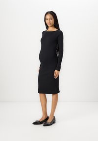 Robe noire à manches longues côtelées avec une coupe ajustée et un col en rond, associée à des chaussures noires ornées d'accents matériels décoratifs.
