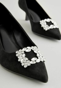 Svarta mockaskor med hög klack och spetsiga tår, med en rhinestone-dekoration i blomsterdesign på framdelen.