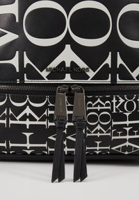 Sac Michael Kors noir et blanc avec un logo imprimé en gras et deux fermetures éclair en métal avec des tirettes en cuir noir sur la poche avant.