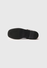 Suela negra de un zapato plano exhibida con la parte inferior hacia arriba, mostrando patrones de agarre texturizados y un logo rectangular cerca del talón.