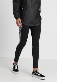 Svarta leggings med en åtsittande design, som har ett kontrasterande abstrakt vitt mönster på sidan. Bärs med svarta och vita sneakers.