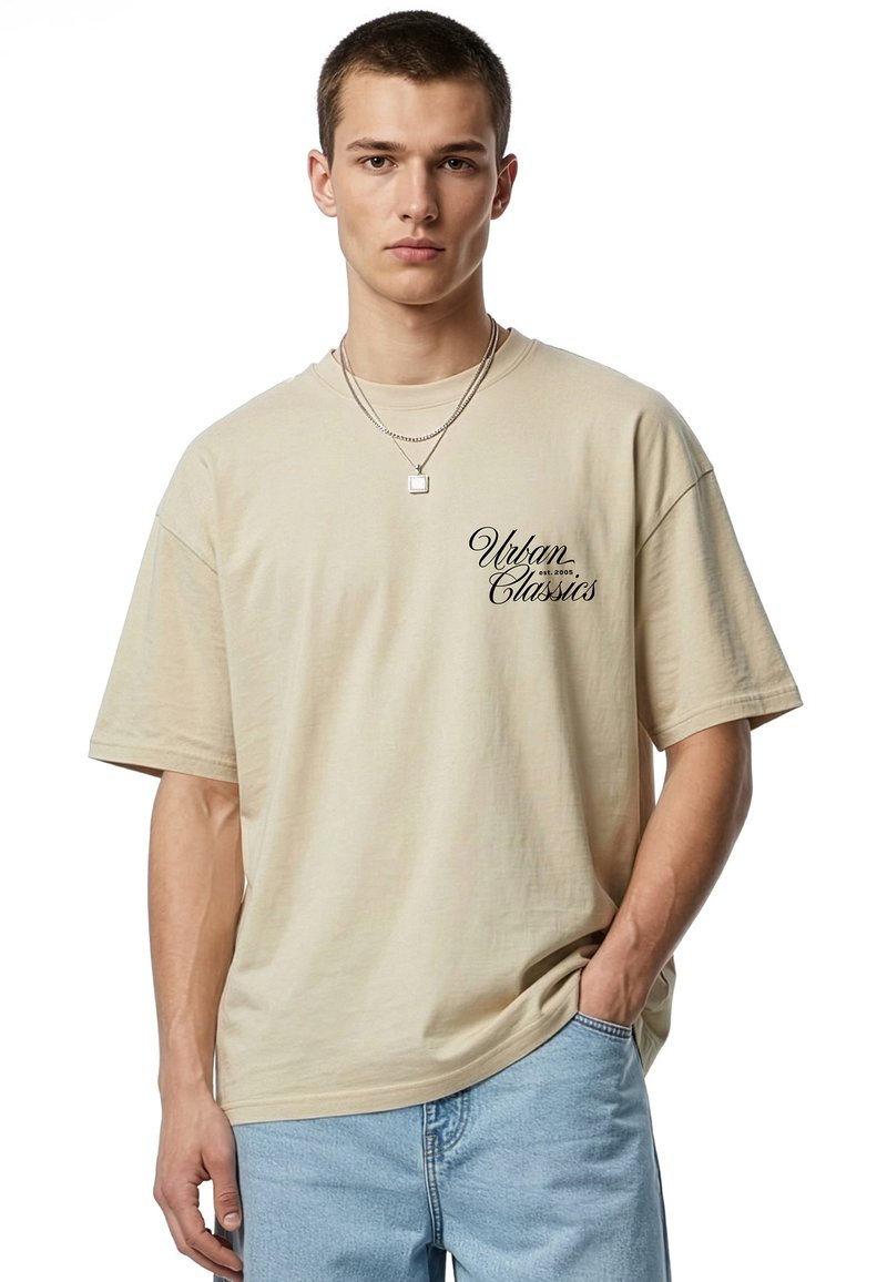 Jeune homme portant un t-shirt beige oversize avec le texte « Urban Classics », un jean bleu clair et deux colliers en argent, posant avec une main dans la poche.